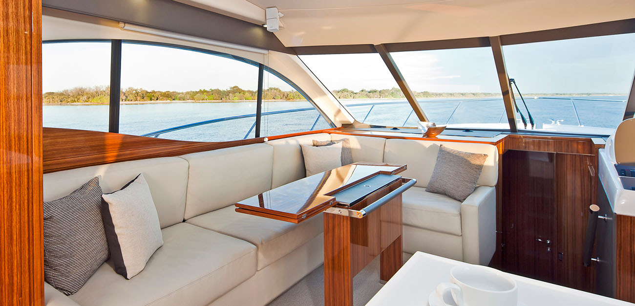 L-shaped cream sofa with gray and white pillows in a wood-paneled luxury yacht lounge, curved windows showing calm water outside.