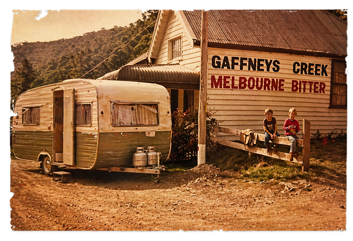 Vintage caravan and trailer parked beside a weathered wooden building with a sign reading 'Gaffney's Creek Melbourne Bitter'; two children sit on a fence watching the scene.