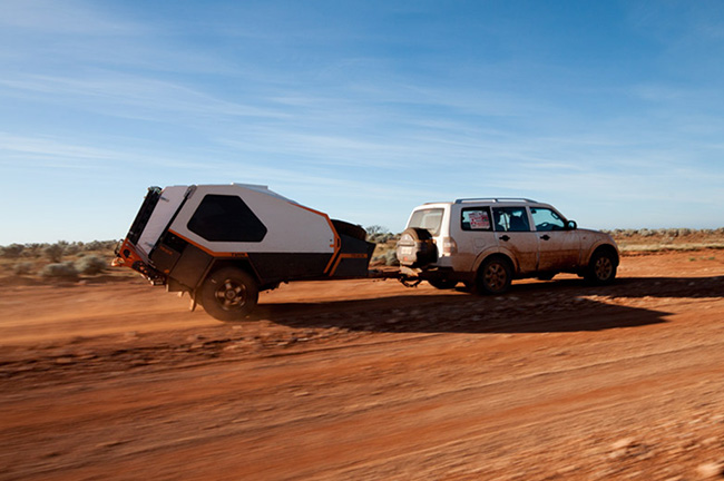 White SUV towing a large off-road camper trailer on a dusty desert road under a blue sky